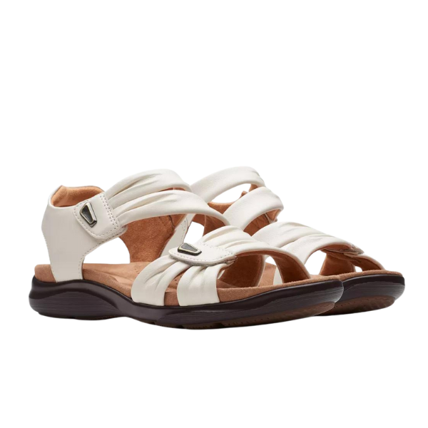 A pair of CLARKS Womens Wide Kitly Ave White sandals with adjustable white straps, Velcro closures, Contour Cushion footbed, cushioned tan insoles, and black rubber soles is displayed on a white background.