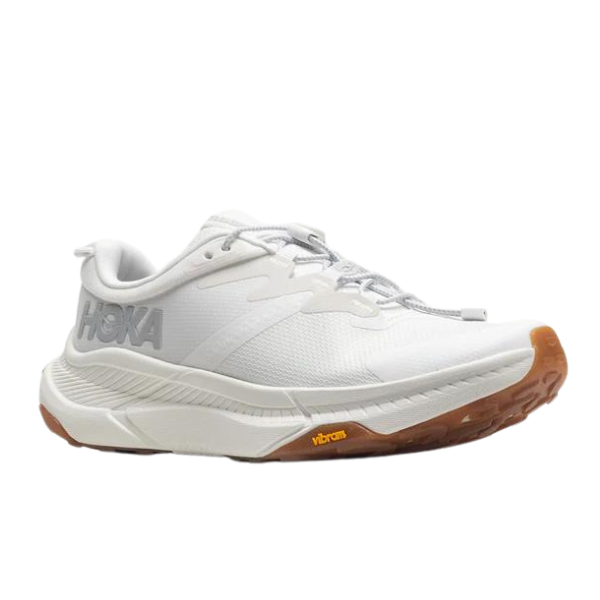 The HOKA Womens Transport White is an athletic sneaker with a thick sole, gray HOKA branding, and a gum rubber outsole featuring a yellow Vibram logo, designed for foot pain relief and shown on a white background.