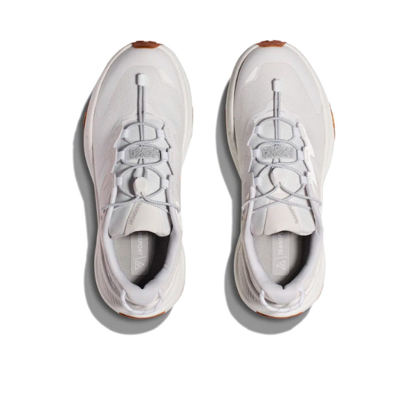 The HOKA Womens Transport White sneakers feature orange-brown soles and elastic loop lacing, viewed from above on a white background. Inspired by HOKA Transport, these sustainable shoes display branded tongue tags.