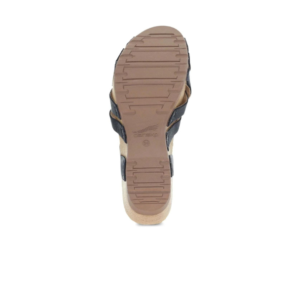 The image shows the bottom of the Dansko Womens Tinley Sandal Black, featuring a tan rubber sole with black straps, visible tread patterns, and the DANSKO logo. The sole also highlights Natural Arch technology for enhanced comfort.