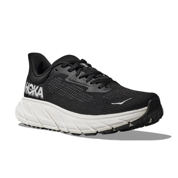 The HOKA Mens Arahi 7 Wide in Black/White features a cushioned white sole, black uppers with black laces, bold white HOKA branding, and J-Frame stability technology. The shoe is angled to show side and front views.