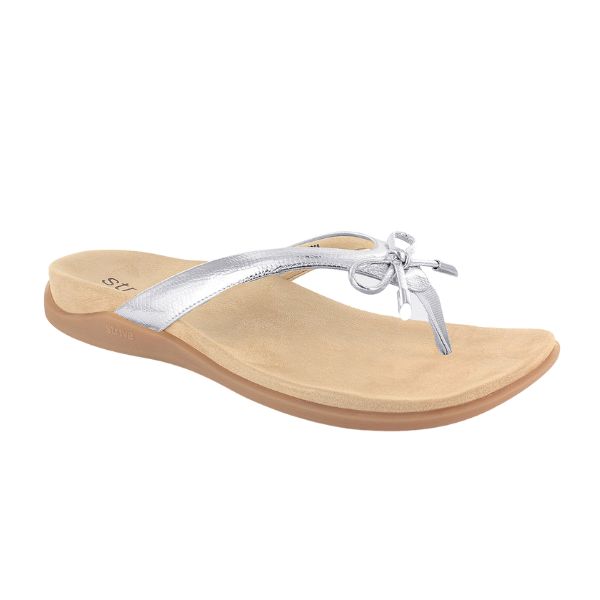 The STRIVE Strive Womens Island Silver is a tan flip-flop sandal with a cushioned sole, subtle arch support, and metallic silver thong straps with a small bow for a feminine touch.