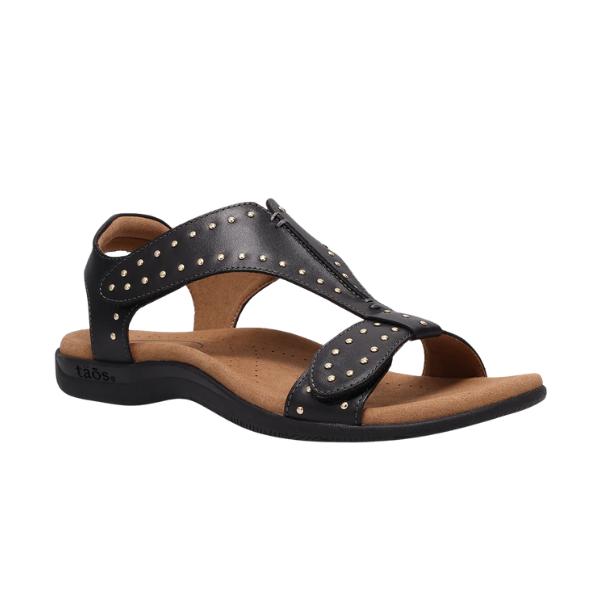 The TAOS Womens Show Stopper Black sandal features a cushioned tan footbed, open toe, ankle strap, gold studded straps, Soft Support technology for comfort, and displays the taös logo on the heel.
