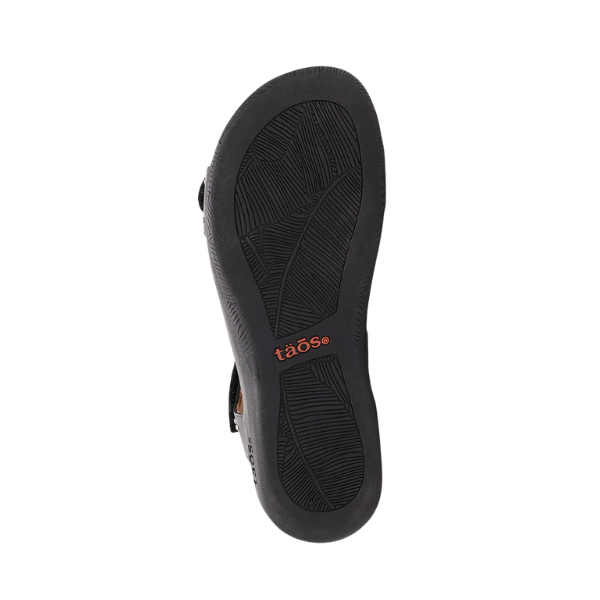 The image displays the black rubber sole of the TAOS Womens Show Stopper Black sandal, featuring textured tread patterns and an orange TAOS logo near the center, set against a white background.