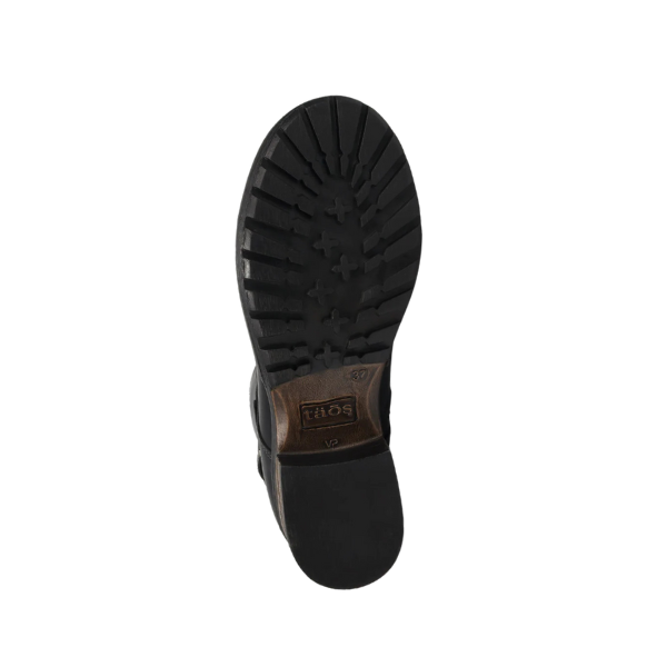 The image shows the bottom view of the Taos Womens Crave Boot in black, featuring a textured rubber sole with deep treads and cross-shaped patterns for traction. A brown TAOS brand label is visible near the heel, emphasizing quality and arch support.