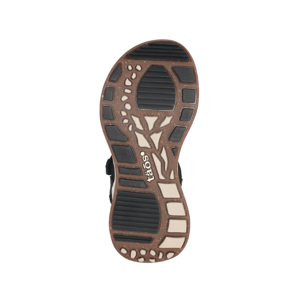 The image displays the sole of the Taos Womens Super Side Black sandal, featuring a textured brown and black pattern with TAOS branding at the center. Its all-terrain rubber outsole includes rectangular and organic shapes for added grip.