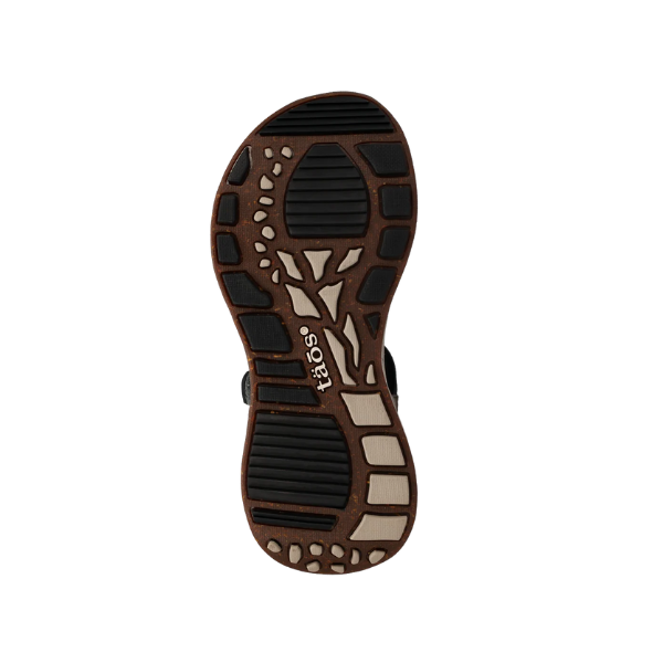 The image shows the sole of the Taos Womens Super Side Dove Multi sandal with a patterned tread in black, brown, and beige. The TAOS brand name appears in white near the center, highlighting its supportive footbed for outdoor use.