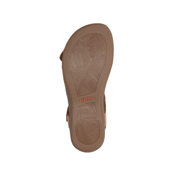 The image shows the sole of the Taos Womens The Show Caramel sandal, made of brown leather with textured patterns and TAOS embossed in orange near the heel. This sandal offers reliable arch support. The background is white.