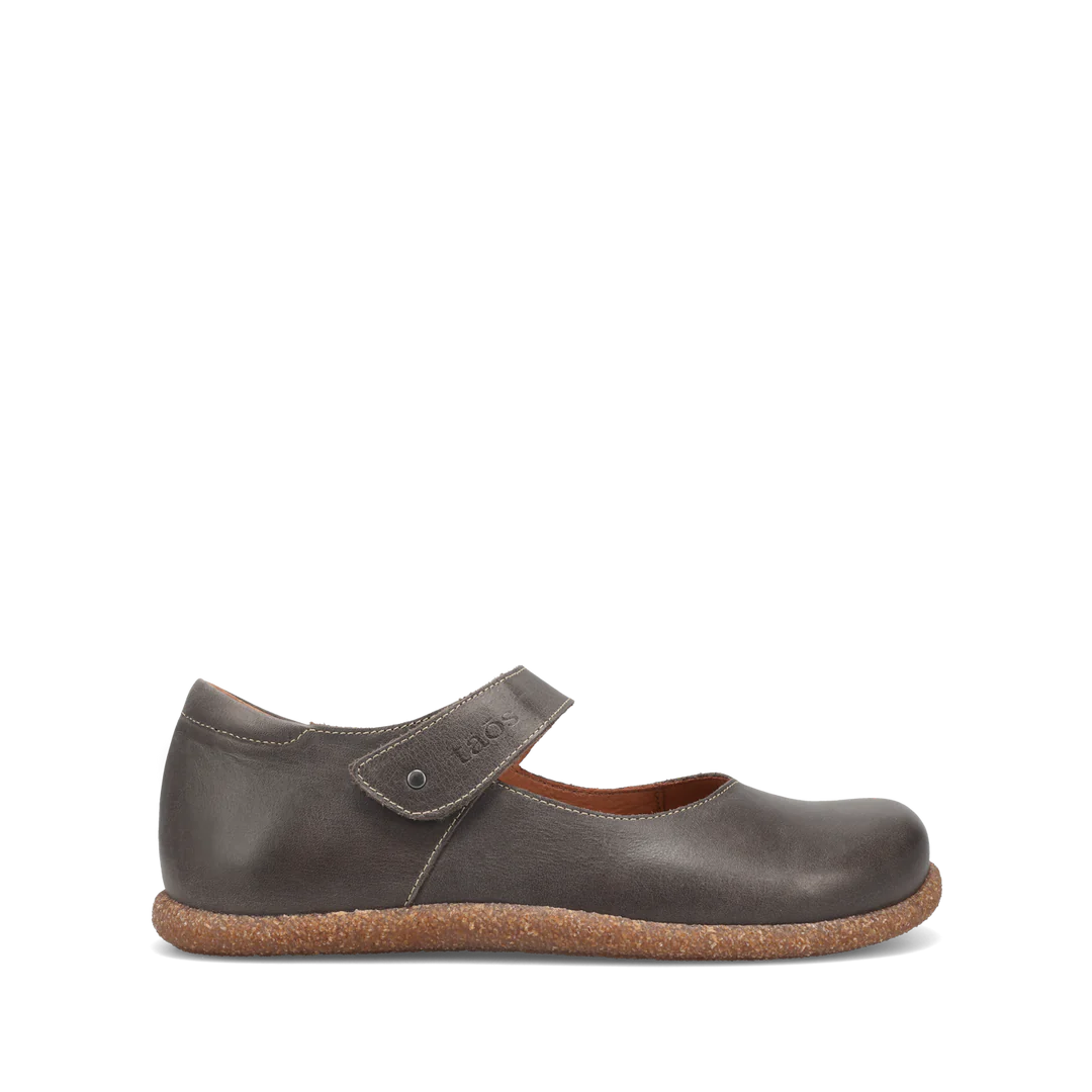 The TAOS Womens Ultimate Grey shoe is a single dark grey Mary Jane with a Velcro strap, rounded toe, and flat textured light brown sole, shown in side view against a plain white background. Made from European leather.