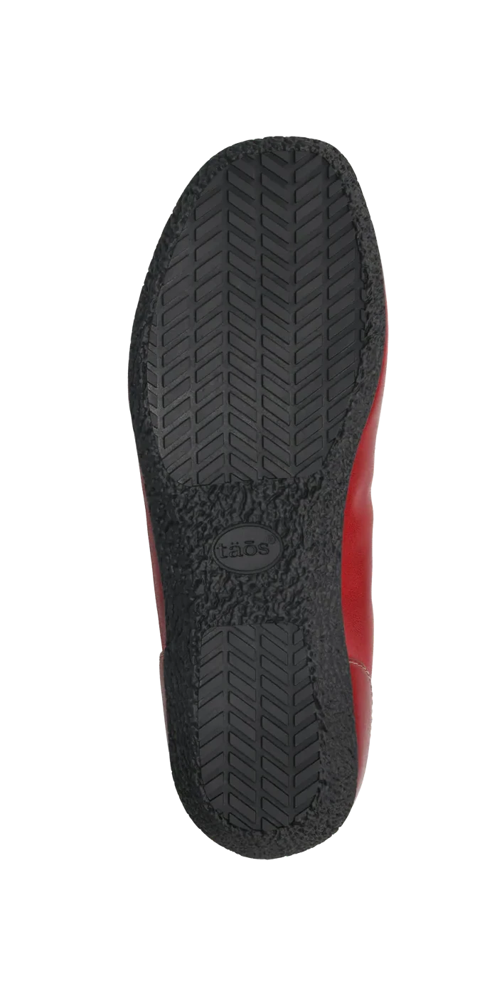 The image shows the black rubber sole of the TAOS Womens Chit Chat Red skimmer flat, featuring a herringbone tread pattern, tide embossed at the center, and a hint of red—ideal for those wanting orthotic-friendly footwear.