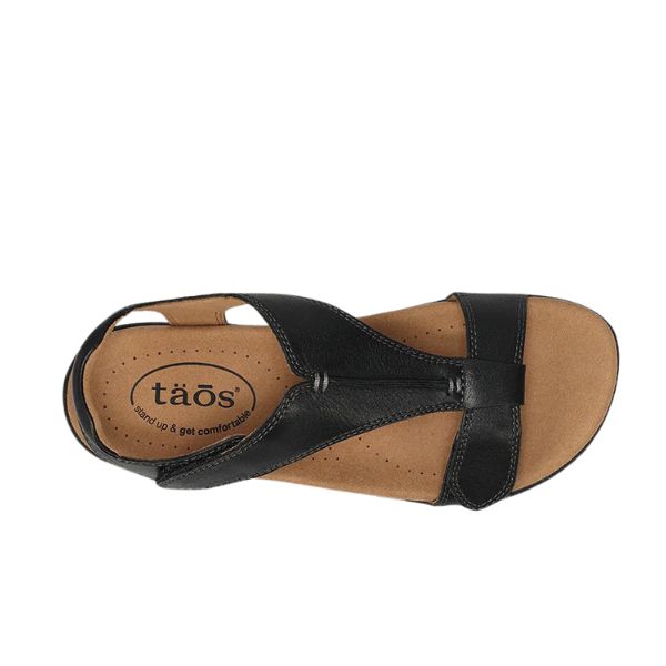 The image features a TAOS Womens The Show Black sandal, a single black strappy leather shoe with a tan cushioned insole and arch support. The “taös” logo is visible on the heel, shown from above against a white background.