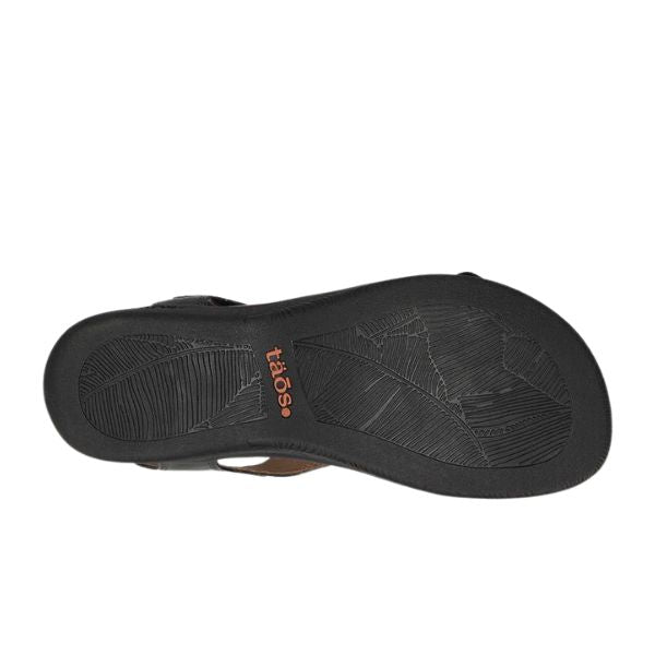 The sole of the Taos Womens The Show Black sandal features textured grip patterns, arch support for all-day comfort, and the TAOS logo in orange printed near the center.