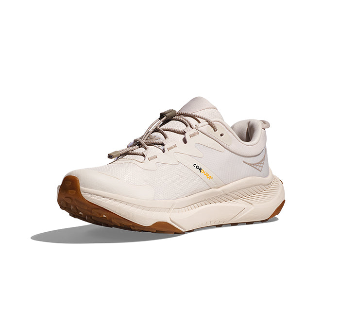 The HOKA Womens Transport Eggnog is a light beige comfort shoe for trail running, made with sustainable materials. It features a rugged brown sole, mesh and synthetic upper, lace-up closure, and HOKA branding—shown on a white background.