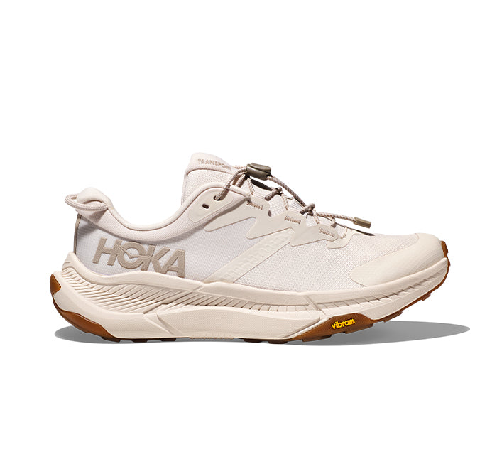 The HOKA Womens Transport Eggnog features a beige mesh upper, white laces, large HOKA logo, cushioned sole, and brown Vibram outsole—crafted with sustainable materials for everyday comfort.