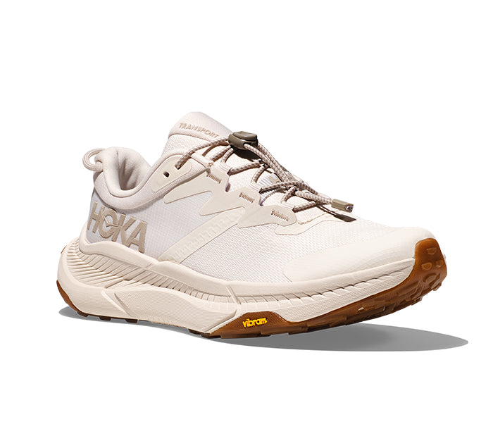 The HOKA Womens Transport Eggnog running shoe, made with sustainable materials, features a white sole, brown Vibram outsole, and toggle laces. Shown at an angle on a white background.