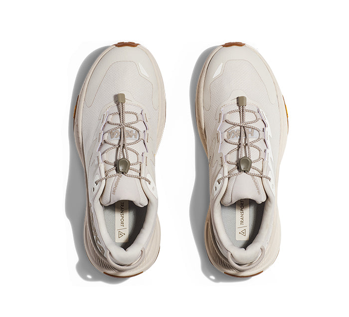 Top view of HOKA Womens Transport Eggnog sneakers by HOKA, featuring light beige color with tan accents, toggle laces, and textured soles—crafted for everyday comfort from sustainable materials, shown side by side on a white background.