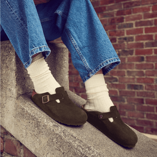 Person wearing brown suede Birkenstock Boston clogs with white socks and blue jeans.