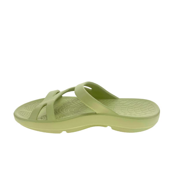 The Sovella Womens PF Green Slide by SOVELLA is a light green slip-on sandal with crisscross straps, a thick sole, arch support, and a textured footbed for comfort and grip. Shown from the side on a white background.