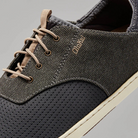 Close-up of the OluKai Men's Nohea Moku Sneaker Grey, a men's casual shoe with textured grey upper, tan laces, visible stitching, cream rubber sole, and "OLUKAI" embroidered in gold on the side.