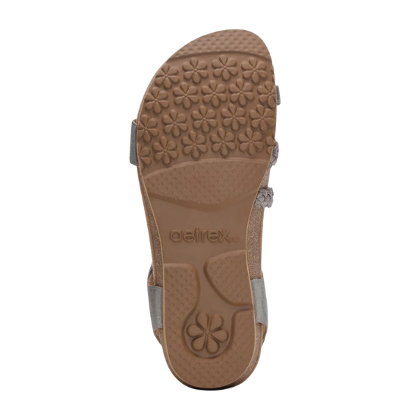 The image shows the bottom view of a brushed-silver Aetrex Womens Jillian sandal sole by AETREX WORLDWIDE INC, featuring a flower-patterned tread, adjustable straps, and Aetrex embossed in the center.