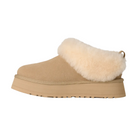 The UGG Women's Tazzelle Mustard Seed slipper features tan suede, a chunky sole, thick light beige faux fur lining, and a pull tab at the back. Shown in side profile against a white background.