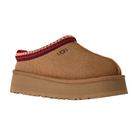 The UGG Women's Tazz II Chestnut slipper features a suede upper, thick light brown sole, red and beige stitching at the opening, soft sheepskin lining, and an embroidered UGG logo on the side.