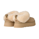 The UGG Women's Tazzelle Mustard Seed slippers feature thick, fluffy sheepskin lining, chunky soles, and back pull tabs—an indoor-outdoor style shown from an angle highlighting the back and side.