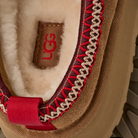 Close-up of the inside heel of an UGG Women's Tazz II Chestnut shoe, showing white sheepskin lining, red and beige stitching, a red UGG logo on a brown tag, and a red fabric pull tab.