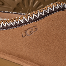 Close-up of the UGG Men's Tasman II Chestnut featuring a tan suede upper, embossed UGG logo, and detailed brown, white, and tan stitching along the edge.
