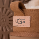 Close-up of the heel of a chestnut UGG Women's Classic Micro boot, showing the stitched UGG logo label. The background is a soft, blurred chestnut suede texture matching this iconic sheepskin footwear.