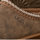 Close-up of the UGG Men's Tasman Baxter Chestnut slipper, featuring a brown suede upper, visible stitching, UGG branding on the side, textured sole details, and cozy UGGplush lining.