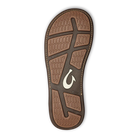 The OLUKAI Mens Tuahine Toffee sandal features a brown sole with textured hexagonal tread, intersecting grip grooves, and Hawaiian-inspired accents, including a central fishhook-shaped logo.