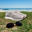 A pair of OLUKAI Mens Ahi Ash/Sterling sneakers and round sunglasses rest on grassy sand by the beach, with waves and a blue sky in the background.