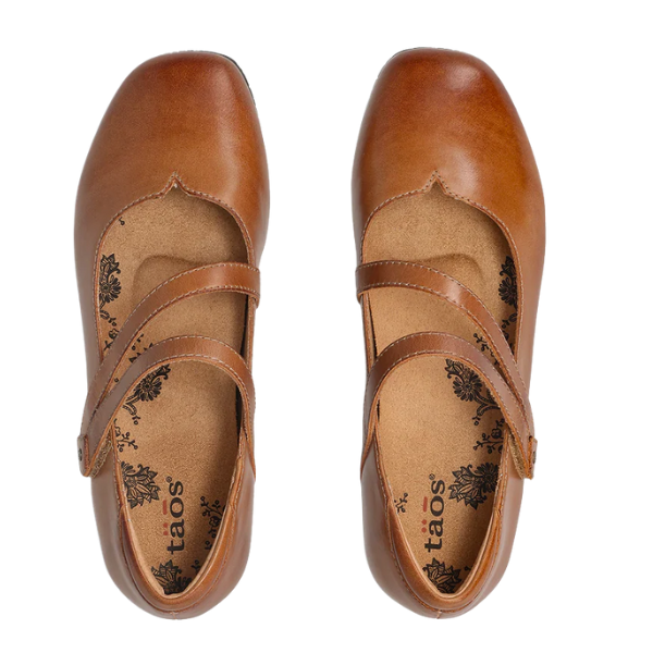 Top view of TAOS Women's Banter Caramel flats featuring floral-patterned insoles, two crisscross straps, and excellent arch support, set against a white background.