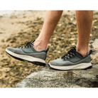 A person wearing OLUKAI Men's Makiki Grey shoes walks on rocky terrain near a sandy, pebbled area, highlighting all-terrain performance.
