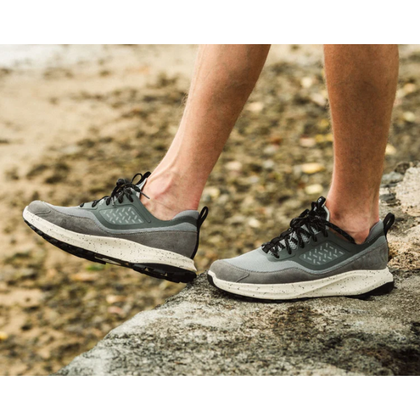 A person wearing OLUKAI Men's Makiki Grey shoes walks on rocky terrain near a sandy, pebbled area, highlighting all-terrain performance.