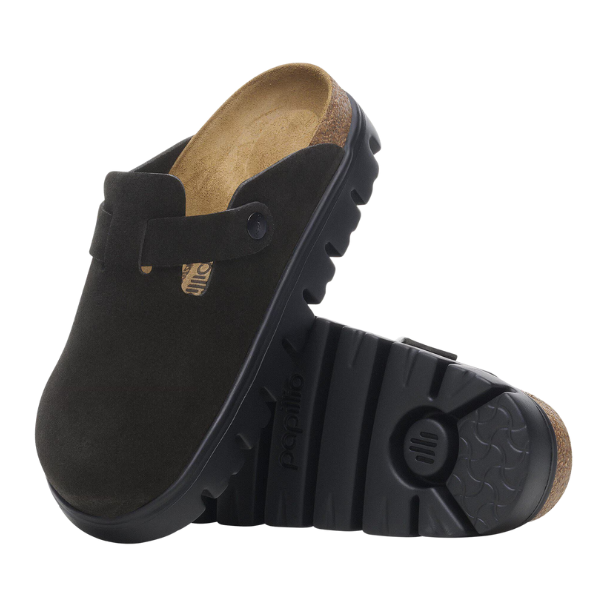 The Birkenstock Women's Boston Chunky Black clogs by BIRKENSTOCK feature black suede uppers, contoured footbeds, and thick, rugged black rubber soles. One shoe stands upright while the other is on its side to display the textured sole.