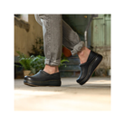 A person in rolled-up gray jeans wears Dansko Women's Platform Pro Cloud Black clogs by DANSKO on a stone floor, with plants and a rustic background—highlighting these stylish, comfortable, and slip-resistant platform clogs.