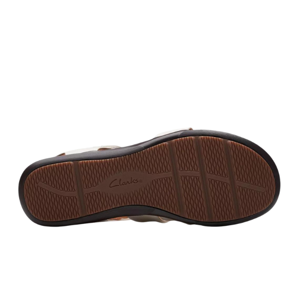 The image shows the sole of a Clarks Womens Wide Kitly Ave White sandal by CLARKS, featuring a textured brown rubber surface with crosshatch tread, the CLARKS logo embossed at the center, and black edges.