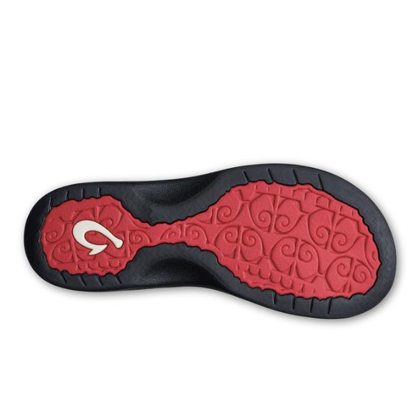 The image shows the sole of the OLUKAI Womens Ohana in Coral/Lehua Flower/Onyx, featuring red and black hues, swirl patterns, an anatomical footbed design, and a white stylized C on the heel.