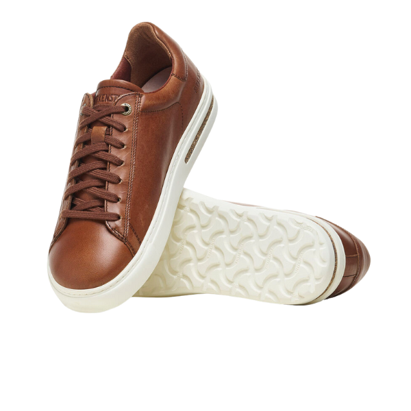 The BIRKENSTOCK Mens Bend Low Cognac features natural brown leather uppers and white textured soles, with one shoe upright and the other on its side to display the contoured footbed for comfort.