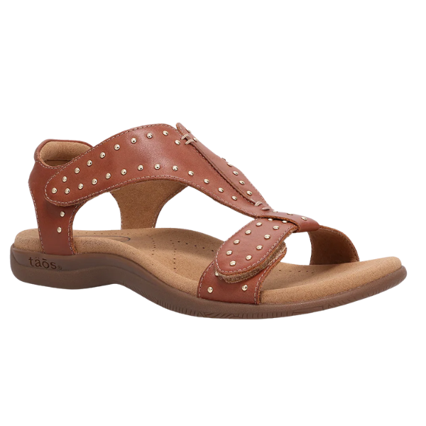 The TAOS Womens Show Stopper Cognac sandal features brown leather, an open toe, Cool Recovery Foam cushioning, decorative stitching, and small metallic studs on the straps, displayed against a white background.