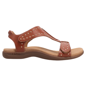 The TAOS Womens Show Stopper Cognac sandal features brown leather, an adjustable ankle strap with gold studs, a cushioned tan Soft Support footbed, a low rubber sole, and embossed TAOS branding on the heel.