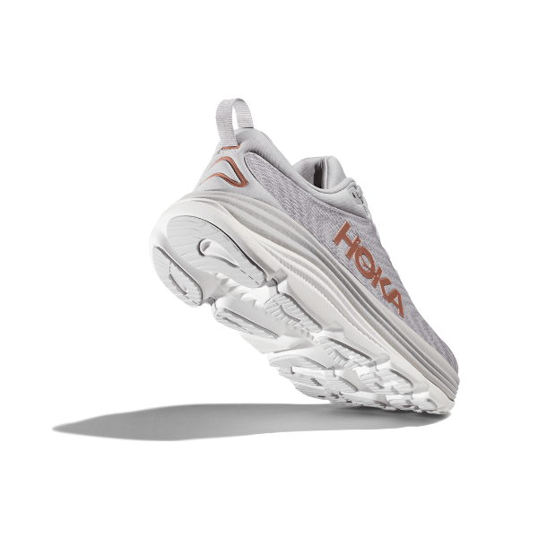 The HOKA Womens Gaviota 5 Wide in Harbor Mist/Rose Gold is shown from a low, rear angle, featuring the stabilizing H-Frame technology and brand name. The light gray shoe has a white sole, rose gold accents, and casts a shadow beneath it.