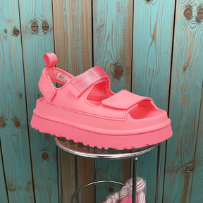 The UGG Womens Goldenglow Sandal Pink, featuring velcro straps and known for comfort, is displayed on a metal stand in front of a turquoise and brown wooden wall.
