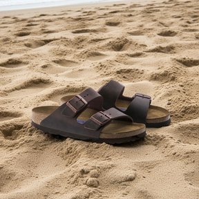 A pair of BIRKENSTOCK Mens Arizona Soft Footbed Oiled Leather Habana sandals, with two buckle straps, rests on sandy beach near the shoreline, ocean visible in the background.