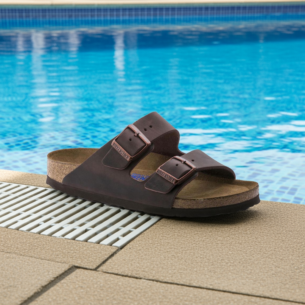 The BIRKENSTOCK Mens Arizona Soft Footbed Oiled Leather Habana sandal rests on the poolside edge, next to a blue-tiled swimming pool.