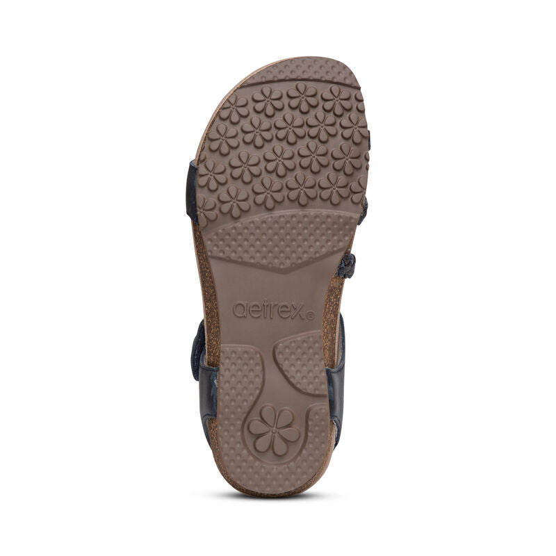 The image shows the sole of the Aetrex Womens Jillian Navy sandal with a textured floral tread, aetrex embossed near the arch, and a brown sole featuring a signature Lynco orthotic footbed.