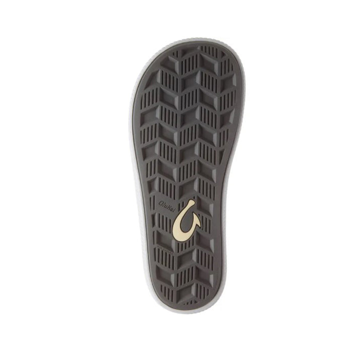 The image shows the bottom of an OluKai Mens Ulele Stone/Stone sandal with a gray geometric tread, yellow hook-shaped logo near the heel, and OluKai branding, highlighting the comfortable design of these mens sandals.