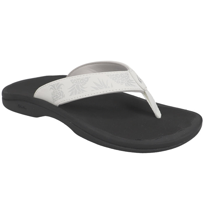 The OluKai Womens Ohana Bright White Hua is a black sandal with a white, floral-patterned strap. Water-resistant and featuring a supportive arch, this flip-flop offers comfort and foot pain relief.